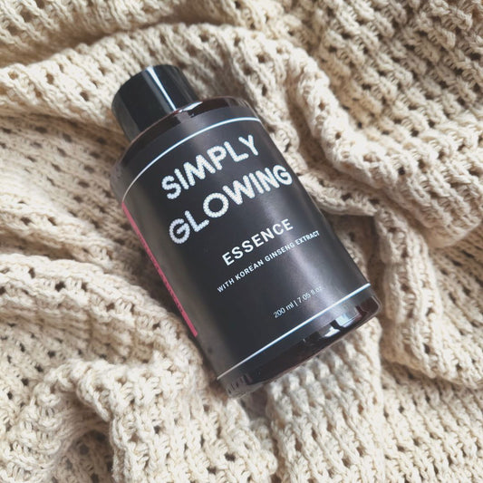 Black bottle labeled 'Simply Glowing Essence' on a textured beige fabric background
